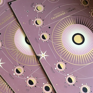 Peut inclure: Une pile de calendriers lunaires roses et dorés avec le texte "Moon Calendar" et "Calendrier Lunaire" imprimé en haut. Chaque calendrier présente un mois différent avec les phases de la lune illustrées.