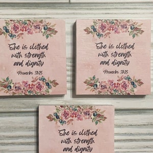 May include: Three pink wooden signs with floral designs and the text "She is clothed with strength and dignity Proverbs 31:25"