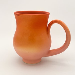 Puede incluir: Una taza de cerámica con cuerpo redondeado y asa curva. La taza es un degradado de naranja y rosa.
