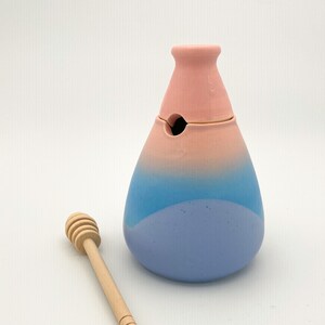 May include: A blue and pink ceramic honey pot with a wooden honey dipper. The pot has a gradient design with a light pink top and a blue bottom.