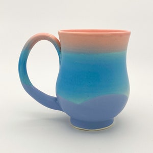 May include: A ceramic mug with a light blue, turquoise, and pink ombre glaze. The mug has a rounded body and a curved handle.