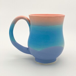 May include: A ceramic mug with a light blue, turquoise, and pink ombre glaze. The mug has a wide, rounded base and a curved handle.