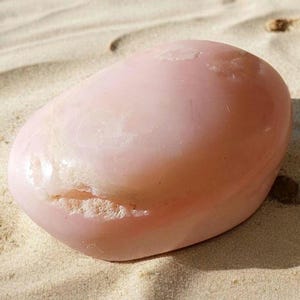 Puede incluir: Una piedra lisa de color rosa, de forma ovalada, con un interior blanco nublado. La piedra está apoyada sobre una superficie arenosa.