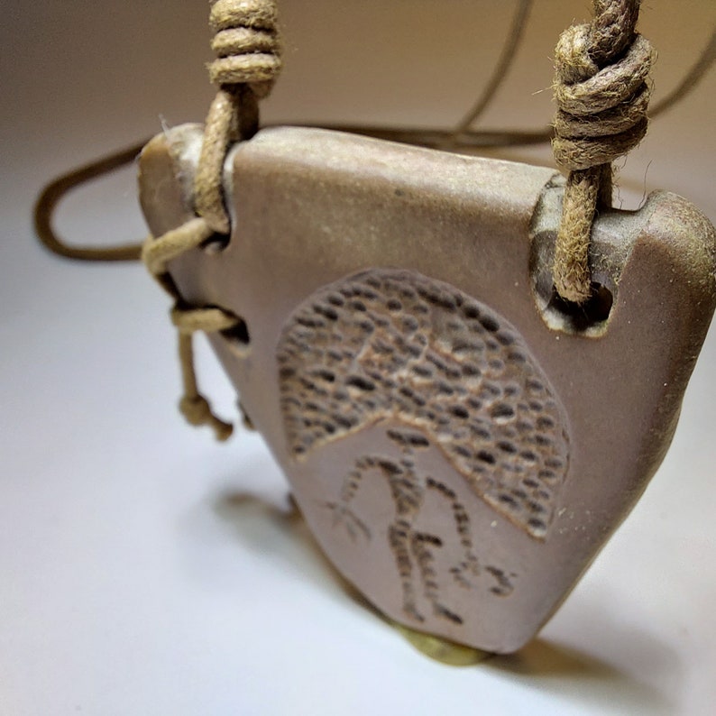 May include: A brown ceramic pendant with a carved design of a person standing under a tree. The pendant is attached to a brown cord.