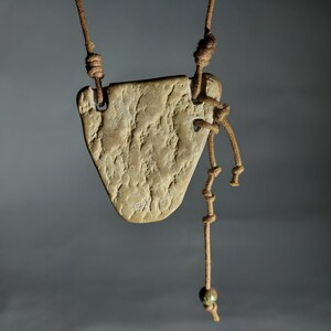 May include: A brown leather cord necklace with a light brown, textured, heart-shaped pendant. The pendant has a small, engraved inscription. The necklace has a small, green bead at the end.