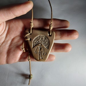 May include: A brown leather cord necklace with a triangular pendant featuring a carved design of a person standing under a mushroom.
