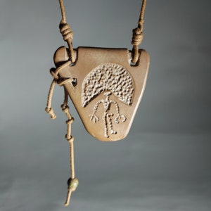 May include: A brown clay pendant with a carved design of a person standing under a tree. The pendant is attached to a brown leather cord with a green bead.