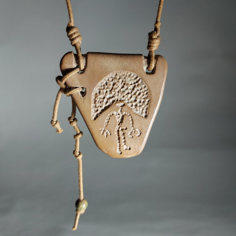 May include: A brown clay pendant with a carved design of a person standing under a tree. The pendant is attached to a brown leather cord with a green bead.