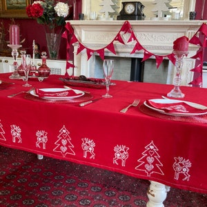 May include: A red tablecloth with a white embroidered design of Christmas trees and reindeer. The tablecloth is set with two place settings, each with a red napkin, a white plate, and a silver fork. The tablecloth is on a table with a white base and a red carpet underneath.