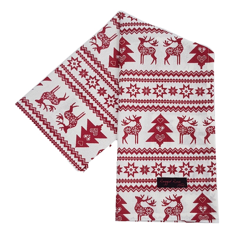 May include: A white and red Christmas table runner with a festive pattern. The design features reindeer, Christmas trees, and star motifs. The runner is made of fabric and has a rectangular shape. The brand name "Ragged Rose" is visible on a black label.