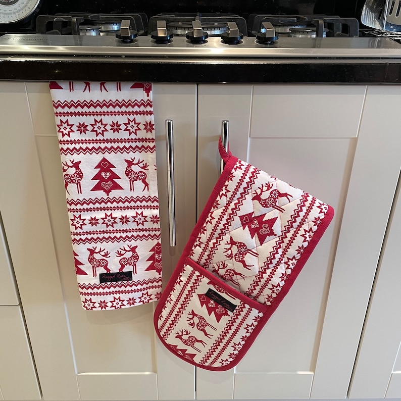 May include: A white and red Christmas kitchen set featuring a patterned design with reindeer, trees, and stars. The set includes a hanging towel and a double oven mitt, both with a red border and a label that reads "Serged Rose".