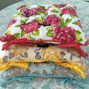 May include: Stack of three square seat cushions with floral patterns. The top cushion has a white background with large red roses and green leaves. The middle cushion has a yellow background with yellow roses. The bottom cushion has a light blue background with gray roses.