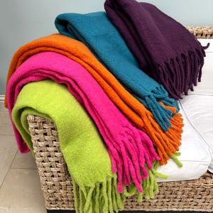 Op de afbeelding: Een verzameling kleurrijke dekens met franjes in limoengroen, felroze, oranje, blauwgroen en paars. De dekens zijn opgevouwen en gestapeld op een rieten stoel, waarbij hun zachte textuur en levendige kleuren worden getoond.