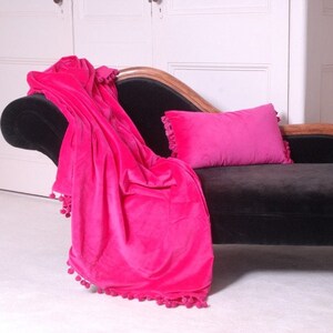 Pu&ograve; includere: Una coperta in velluto rosa brillante con pompon su tutti i bordi &egrave; drappeggiata su una chaise longue nera. Un cuscino in velluto rosa abbinato &egrave; sulla chaise longue.