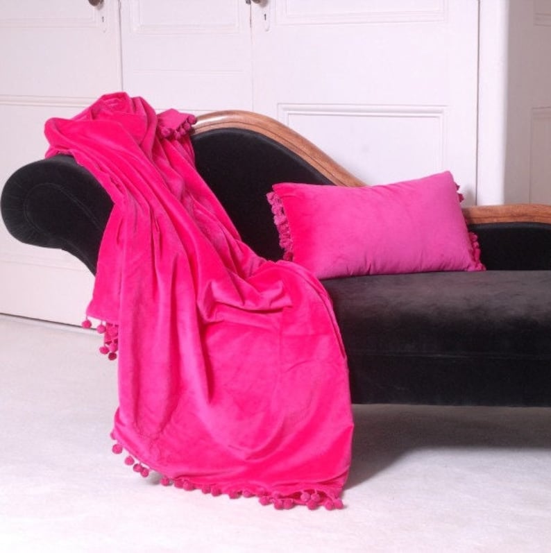 Pu&ograve; includere: Una coperta in velluto rosa brillante con pompon su tutti i bordi &egrave; drappeggiata su una chaise longue nera. Un cuscino in velluto rosa abbinato &egrave; sulla chaise longue.