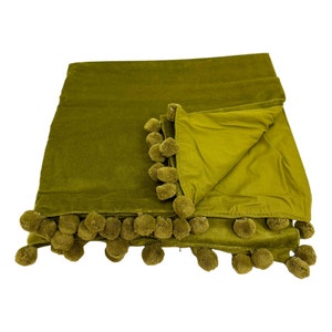 May include: A green velvet throw blanket with a pom-pom trim. The blanket is folded in half and the pom-poms are visible on both sides.
