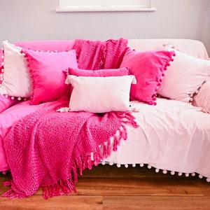 Pu&ograve; includere: Un divano rosa con un bordo bianco a pompon, coperto da una coperta di maglia rosa e diversi cuscini rosa.