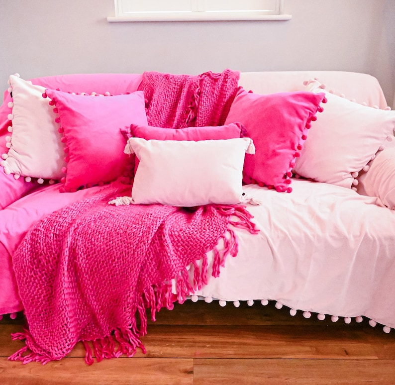 Pu&ograve; includere: Un divano rosa con un bordo bianco a pompon, coperto da una coperta di maglia rosa e diversi cuscini rosa.