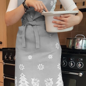 May include: A gray Christmas-themed apron with white snowflakes, reindeer, and Christmas trees. The apron has a large pocket in the front.