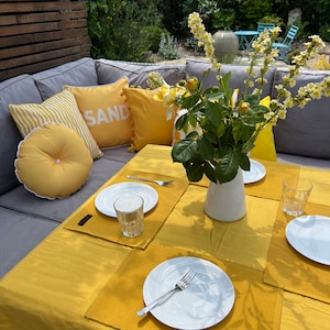 Könnte beinhalten: Eine gelbe Tischdecke mit vier Gedecken, jedes mit einem weißen Teller, einer Gabel und einem Glas. Es gibt gelbe Tischsets mit einer gewebten Textur. Eine Vase mit gelben Blumen steht in der Mitte des Tisches. Der Tisch ist in einem Garten mit einem grauen Ecksofa und Kissen mit der Aufschrift "SAND" gedeckt.