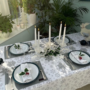 May include: A white tablecloth with a snowflake pattern is set for a festive dinner. The table is decorated with a silver candelabra with four white candles, green holly sprigs, and white plates with green holly sprigs.