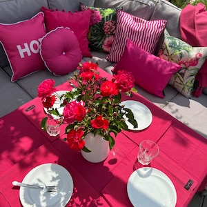 Op de afbeelding: Buitentafel met een magenta tafelkleed, witte borden en glazen. Een witte vaas bevat rode rozen. Kussens in roze tinten en kussens met patronen sieren de zithoek. Het woord "HOT" staat op een kussen.
