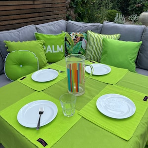 May include: A green tablecloth with four place settings, each with a white plate, a silver fork, and a green placemat. There is a glass of water and a jug of colourful striped water on the table. The table is set in a garden with a grey sectional sofa with green and white cushions.