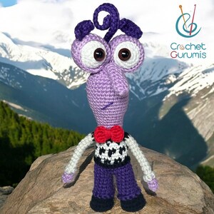 Puede incluir: Juguete amigurumi de crochet morado con una corbata de lazo roja, un chaleco a cuadros blanco y negro y pantalones morados. El juguete tiene grandes ojos blancos y un mechón de pelo rizado morado. El juguete está de pie sobre una roca con un fondo de montaña.