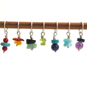 Könnte beinhalten: Ein Set aus zehn bunten Edelstein-Maschenmarkierern, jeder mit einer anderen Farbe und Form. Die Markierer sind auf einer Holzleiste aufgereiht.