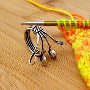 Op de afbeelding: Een zilveren brei-instrument met een uniek ontwerp, naast een breinaald en een stuk gebreide stof in geel en oranje. Het garen is geel en oranje. Het gereedschap heeft een ring en drie gebogen armen.