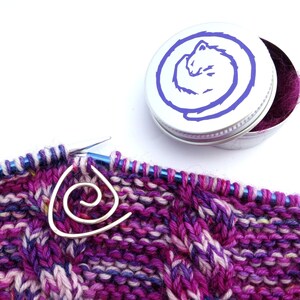 May include: A purple and white knitted fabric with a silver spiral stitch marker. The marker is in the shape of a triangle with a spiral in the centre. A small silver tin with a purple curled cat design is in the background.