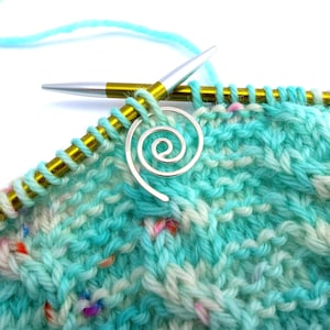 May include: A close-up of a turquoise and white knitted fabric with a silver spiral stitch marker and a pair of gold knitting needles.