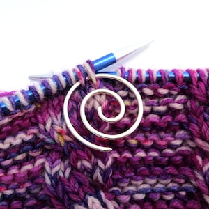 Pode incluir: Um close-up de um projeto de tricô em andamento, com um fio roxo e rosa com um marcador de ponto em espiral prateado. As agulhas de tricô são azuis e brancas.