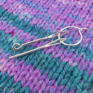 May include: A silver knitting needle with a hook on one end and a loop on the other, resting on a purple and blue knitted fabric.
