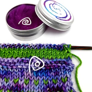 Puede incluir: Primer plano de un proyecto de tejido con hilo verde y morado. Un marcador de punto plateado en forma de espiral está unido al tejido. Un pequeño bote con tapa morada y una etiqueta blanca con un diseño colorido está en el fondo.