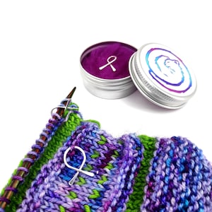 Könnte beinhalten: Nahaufnahme eines Strickprojekts mit grünem, violettem und blauem Garn. Zwei silberne Maschenmarkierer befinden sich auf den Stricknadeln. Im Hintergrund befindet sich eine kleine Dose mit einem violetten Deckel und einem silbernen Banddesign.