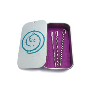 May include: A silver tin with a teal cat design on the lid. The tin contains three silver cleaning tools, two straight and one twisted.