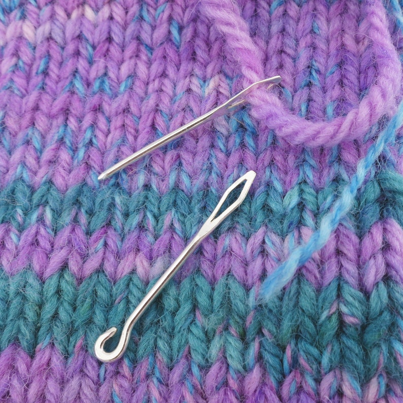May include: Two silver knitting needles on a purple and blue knitted fabric.