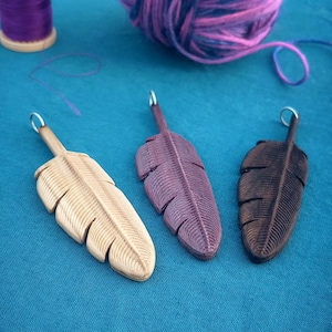 Peut inclure: Trois breloques en bois en forme de plume de différentes couleurs : bois naturel, violet et marron. Les plumes présentent un motif sculpté détaillé et sont suspendues à des anneaux argentés.