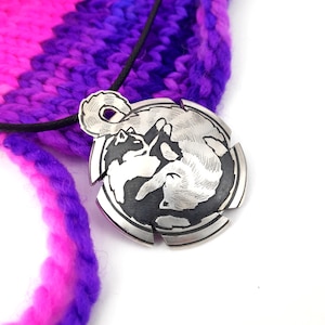 Könnte beinhalten: Eine silberne Halskette mit einem Anhänger, der ein detailliertes, graviertes Design von zwei ineinander verschlungenen Katzen zeigt. Der Anhänger ist rund mit einem strukturierten Rand und hängt an einer schwarzen Schnur.