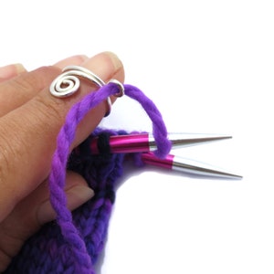May include: A close-up of a person's hand using a silver knitting ring to hold a purple yarn while knitting with two knitting needles. One needle is pink and the other is silver.