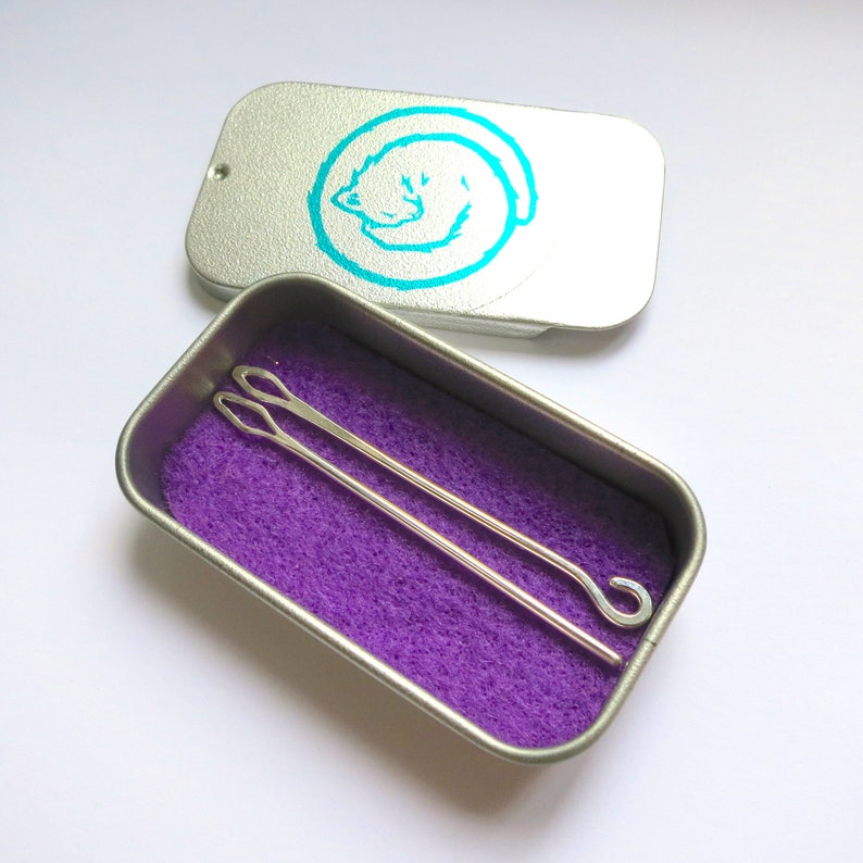 May include: Two silver crochet hooks in a small tin with a purple interior. The tin has a turquoise cat design on the lid.