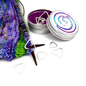 Puede incluir: Primer plano de un proyecto de tejido con una aguja de tejer de madera y un pequeño recipiente de hojalata con un interior morado. El recipiente de hojalata tiene una tapa plateada con un gráfico colorido de un remolino y un oso. Hay varios marcadores de punto triangulares plateados dispersos alrededor del proyecto de tejido.