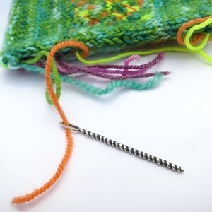 May include: A close-up of a tapestry needle threaded with orange yarn, with a colorful knitted fabric in the background. The needle is silver with black stripes.