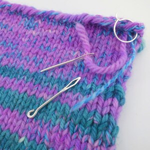 May include: Close-up of a purple and teal knitted fabric with two metal knitting needles and a silver ring. The fabric is a chevron pattern.