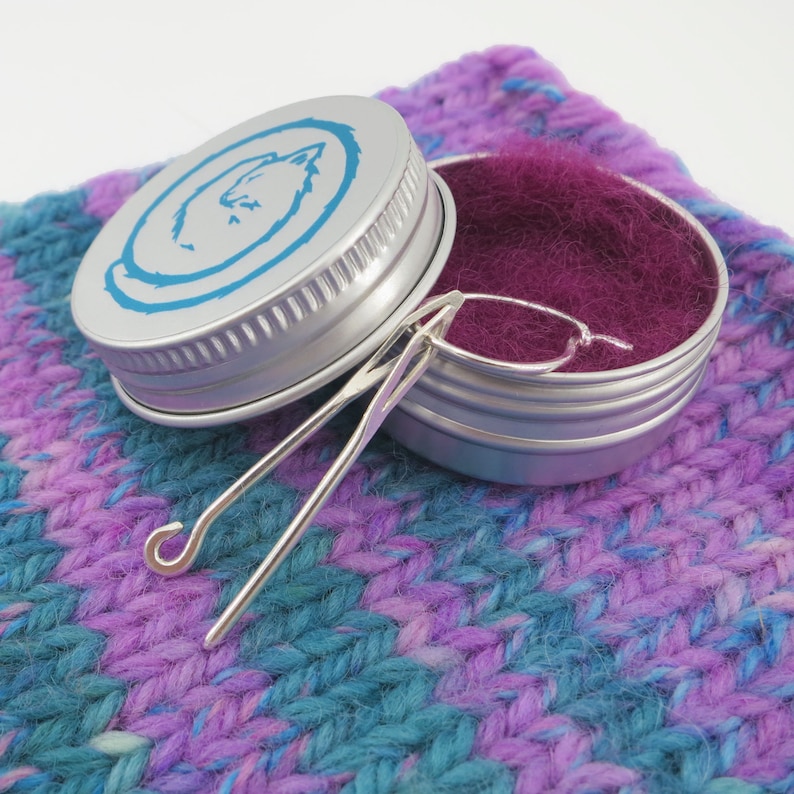 May include: A silver metal container with a blue and white wolf design on the lid. The container is open and contains a small, silver crochet hook and a small amount of purple yarn. The container is sitting on a purple and blue knitted fabric.