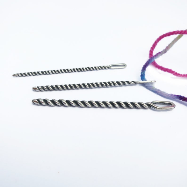 May include: Three silver crochet needles with twisted metal shafts and eye loops. The needles are arranged in order of size, with the largest needle at the bottom and the smallest needle at the top.