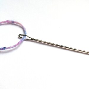 May include: A close-up of a needle threaded with light blue and pink yarn.