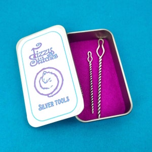 以下が含まれることがあります： 「Fizzy Stitches」と「Silver Tools」の文字が印刷された白いブリキ缶。缶が開いており、中には銀色の針通し2本が入っています。