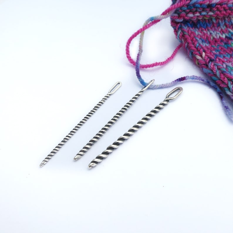 May include: Three silver knitting needles with a twisted design. The needles are different sizes and are arranged in a row on a white surface.